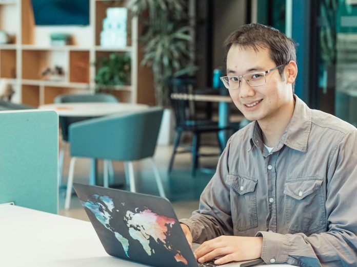 Asian man smiling while working on a laptop in a modern office setting.
